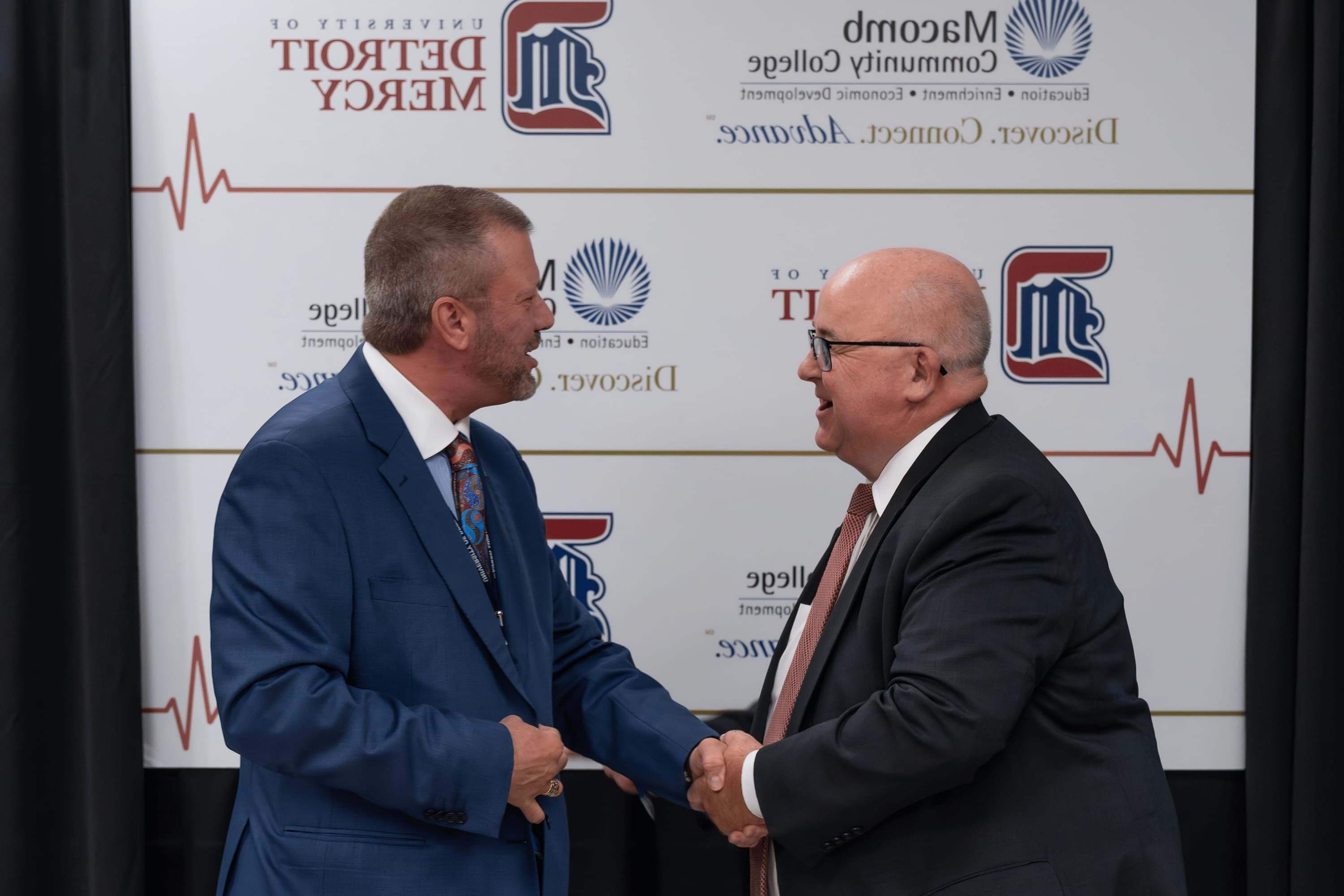 Two men smile and shake hands and look at each other, with logos for University of Detroit Mercy and Macomb Community College on a board behind them.