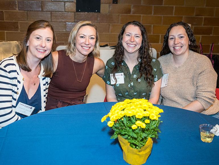 Four women sit around a table and pose for a photo during a Homecoming event. A yellow planted flower is on the table in front of the women.