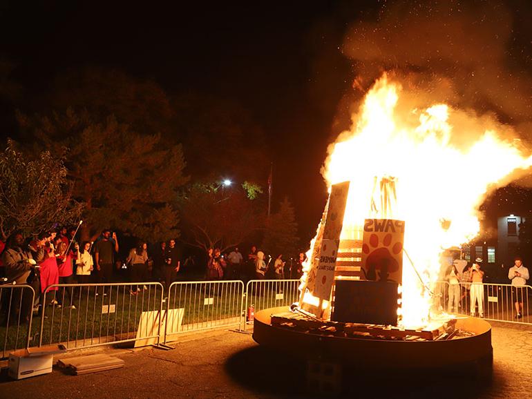 People gather on the grass around a large bonfire in a parking lot. The bonfire features several decorated signs. 