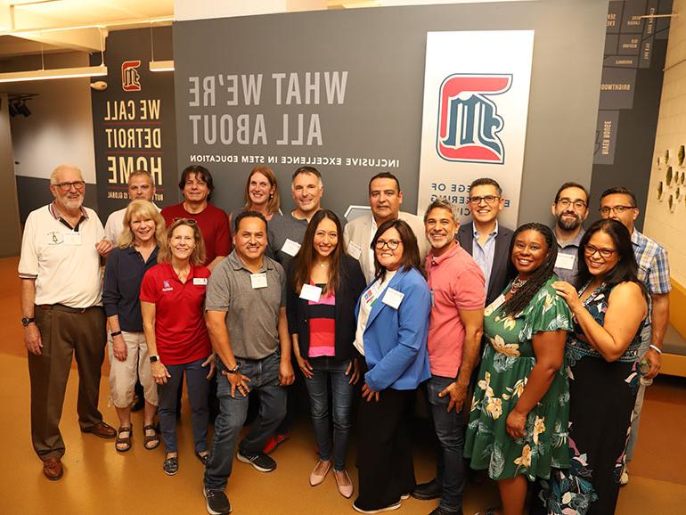 A group of alumni pose for a photo in the College of Engineering & Science's hallway, in front of a decorated wall with several UDM logos and the following text: Detroit Mercy College of Engineering & Science. What we're all about. Inclusive excellence in STEM education. We call Detroit Home. 