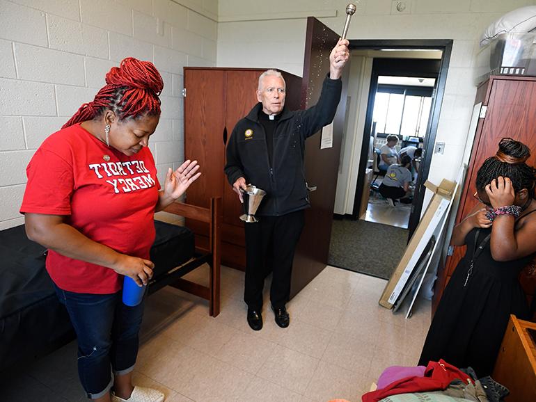 A father blesses a room with two people standing inside of it. One wears a red Detroit Mercy t-shirt. Across the hallway others can be seen in a dorm room of Shiple Hall.