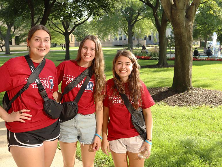Three students wearing red University of Detroit Mercy t-shirts smile for a photo during a sunny day outdoors on the McNichols Campus.