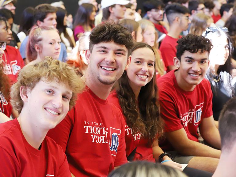 Four students wearing red University of Detroit Mercy t-shirts smile for a photo while sitting indoors. Other students are pictured behind them.