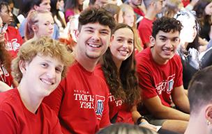 四名学生穿着红色的底特律仁爱大学t恤，坐在室内微笑着拍照, 其他学生在后台.